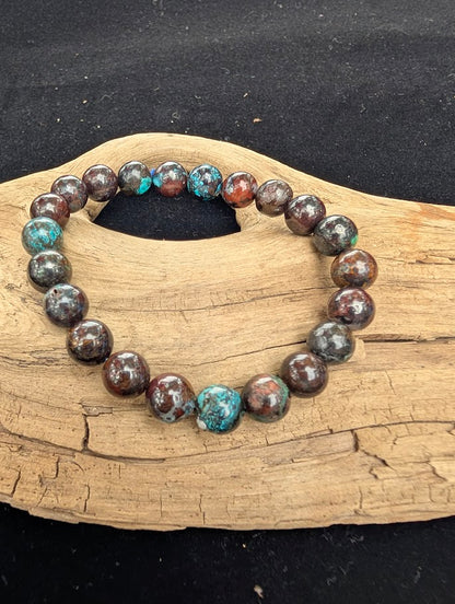 8mm polished Phoenix Stone beaded stretch bracelet resting on a driftwood display, showing a dark copper matrix with teal and green inclusions.