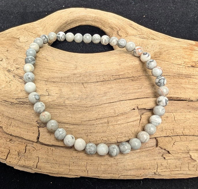 4mm polished Picasso Jasper beaded stretch bracelet resting on a driftwood display, showing a pale grey matrix with abstract dark banding.