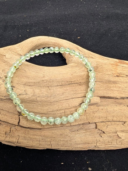 4mm polished Prehnite beaded stretch bracelet resting on a driftwood display, showing pale green translucency and dark Epidote inclusions.