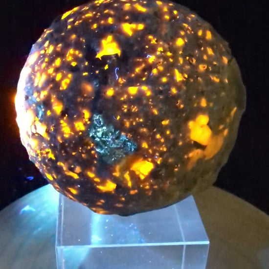 A 98 mm (3.86 in) Yooperlite carved moon sphere glowing bright orange under UV light on an acrylic display stand.