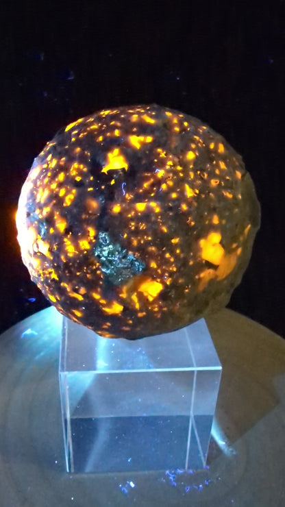 A 98 mm (3.86 in) Yooperlite carved moon sphere glowing bright orange under UV light on an acrylic display stand.