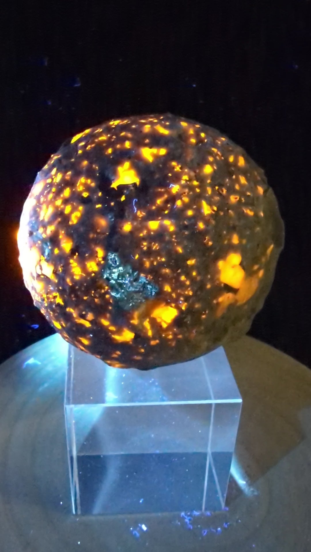 A 98 mm (3.86 in) Yooperlite carved moon sphere glowing bright orange under UV light on an acrylic display stand.