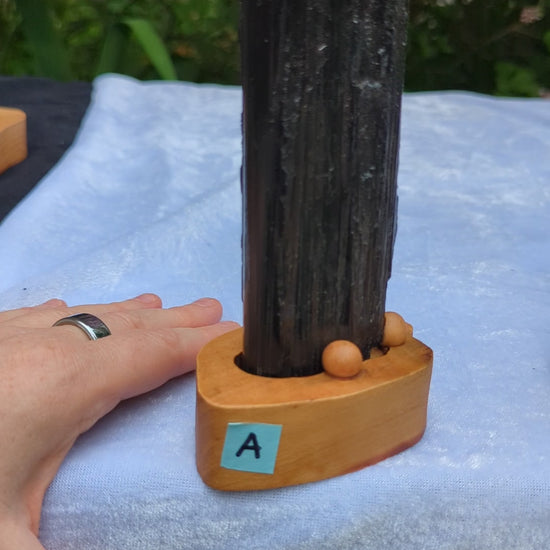 A hand rotates a vertical Black Tourmaline crystal tower mounted on a custom wooden base. The camera pans across a collection of six distinct black tourmaline specimens labeled A through F, highlighting their long, deep, natural vertical striations and high-luster terminations.