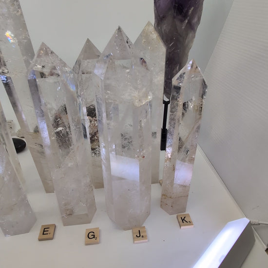 A sweeping video of a collection of polished Clear Quartz towers on a white surface, with a hand picking up and rotating two individual pieces to display their high transparency, internal veiling, and natural golden inclusions.