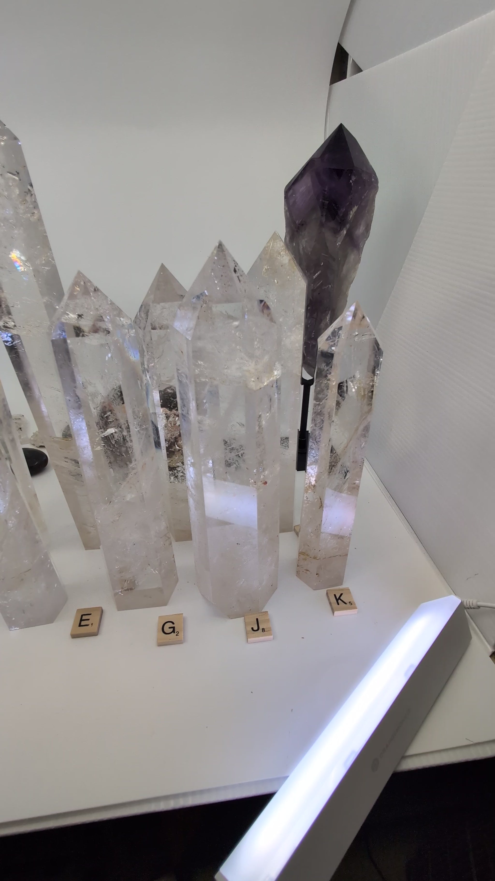 A sweeping video of a collection of polished Clear Quartz towers on a white surface, with a hand picking up and rotating two individual pieces to display their high transparency, internal veiling, and natural golden inclusions.