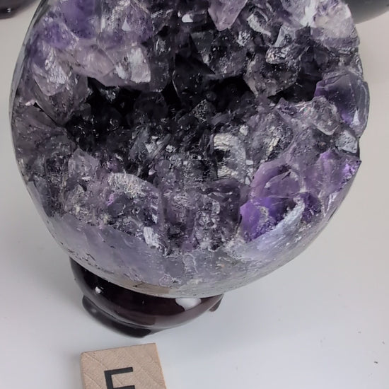 Video showcase of 14 premium Uruguayan Amethyst 'Pac-Man' spheres by Legacy Crystals. The camera pans over specimens A through N, highlighting the deep purple crystal vugs, polished black basalt exteriors, and unique sizes ranging from 2.4 to 3 inches. A hand picks up several spheres to demonstrate the high-gloss polish, internal crystal fire, and scale. The video emphasizes the contrast between the sleek modern rind and the 130-million-year-old amethyst geode interiors.