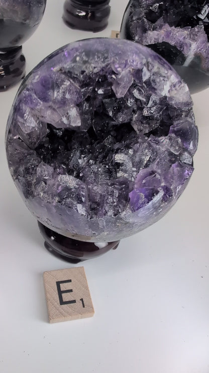Video showcase of 14 premium Uruguayan Amethyst 'Pac-Man' spheres by Legacy Crystals. The camera pans over specimens A through N, highlighting the deep purple crystal vugs, polished black basalt exteriors, and unique sizes ranging from 2.4 to 3 inches. A hand picks up several spheres to demonstrate the high-gloss polish, internal crystal fire, and scale. The video emphasizes the contrast between the sleek modern rind and the 130-million-year-old amethyst geode interiors.
