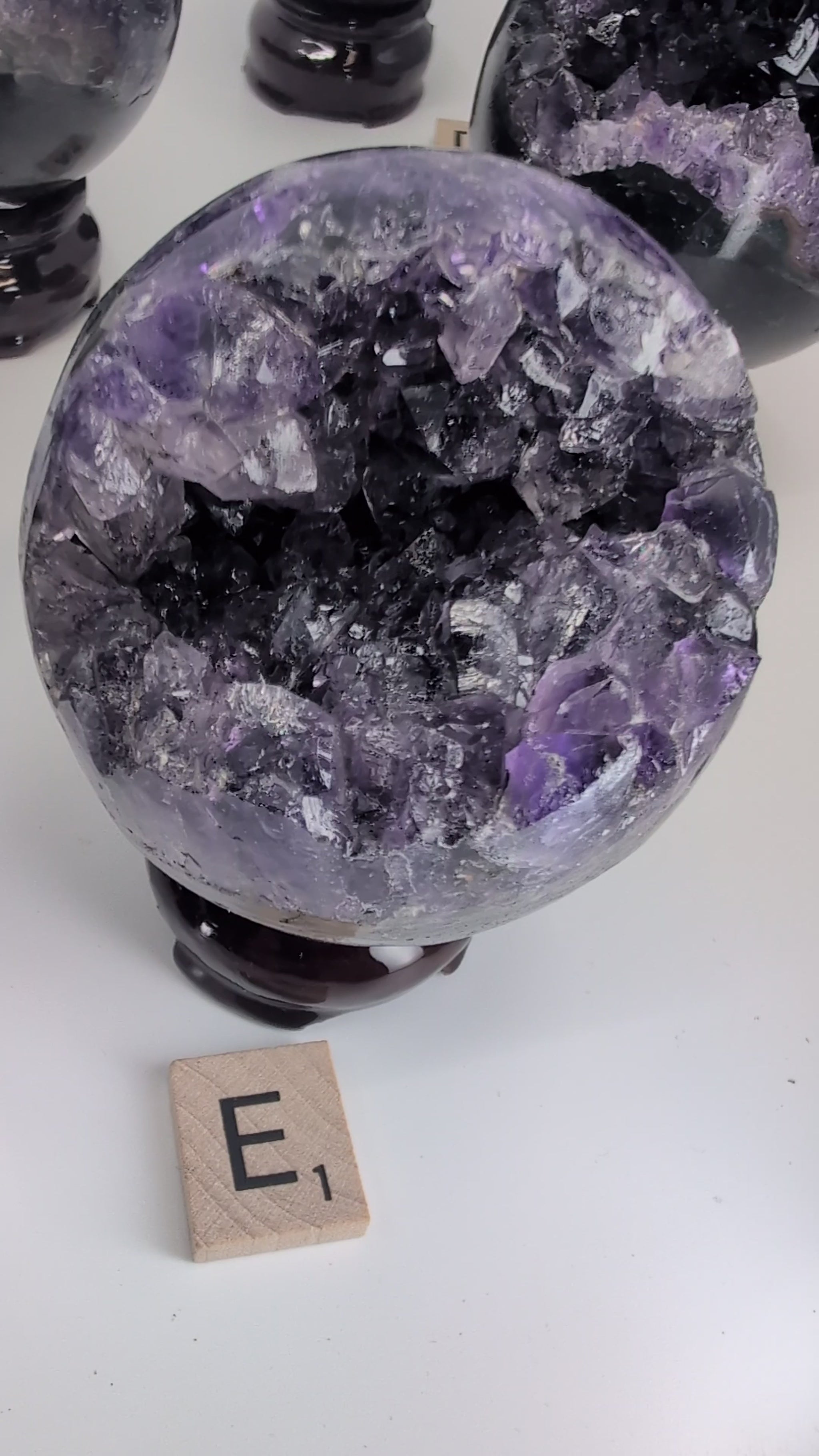 Video showcase of 14 premium Uruguayan Amethyst 'Pac-Man' spheres by Legacy Crystals. The camera pans over specimens A through N, highlighting the deep purple crystal vugs, polished black basalt exteriors, and unique sizes ranging from 2.4 to 3 inches. A hand picks up several spheres to demonstrate the high-gloss polish, internal crystal fire, and scale. The video emphasizes the contrast between the sleek modern rind and the 130-million-year-old amethyst geode interiors.