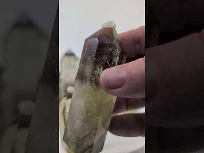 A video tour of nine natural, polished Smoky Citrine towers from Brazil. The camera pans across the standing collection, and a hand picks up several individual pieces to showcase their striking bicolored zoning—from deep smoky quartz bases to pale golden-yellow tips. Close-up shots reveal excellent internal clarity, natural fracture planes creating iridescent rainbows, and dark, floating mineral inclusions suspended within the polished quartz.