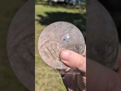 A hand holding a large, 1.12 kg clear Brazilian quartz sphere outdoors in bright natural sunlight. The sphere is slowly rotated to showcase its exceptional glass-like clarity, internal prismatic fracture planes that flash with rainbow iridescence, golden-yellow hematite inclusions, and a large, actively moving water bubble (enhydro) trapped inside a central cavity.