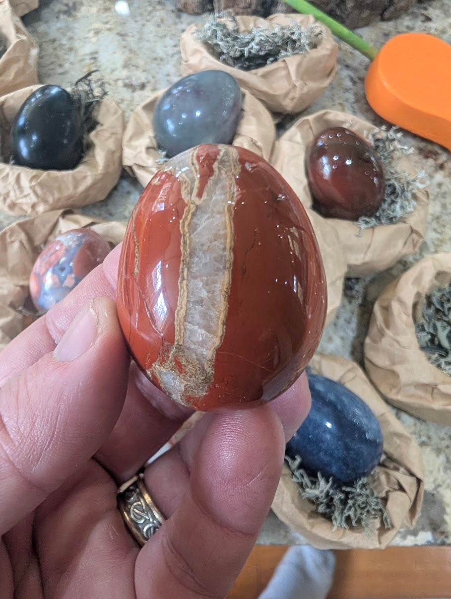 A 2-inch polished Red Jasper egg held in a hand, displaying deep brick-red natural stone intersected by a prominent white quartz vein.