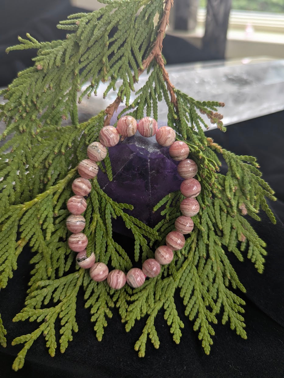 Argentinian Rhodochrosite bracelet draped over a purple amethyst point with green cedar foliage for lifestyle branding.