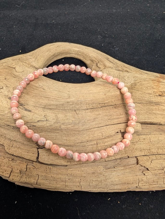 4mm A-grade polished Rhodochrosite beaded stretch bracelet resting on a driftwood display, showing opaque rose-pink tones and prominent white calcite banding.