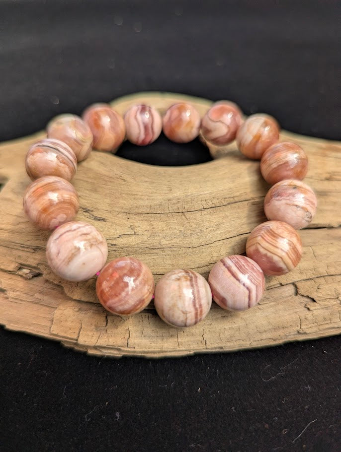 Intuitively selected 14mm polished Argentinian Rhodochrosite beaded stretch bracelet resting on a driftwood display, showcasing highly polished pink and white stalactitic banding.