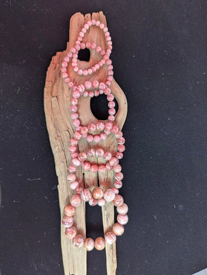 Suite of intuitively selected Argentinian Rhodochrosite beaded stretch bracelets displayed side-by-side on driftwood, showing varied sizes from 6mm to 14mm.