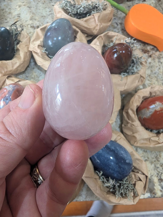 A 2-inch polished Rose Quartz egg held in a hand, displaying soft pink tones, natural translucency, and internal crystal veils.