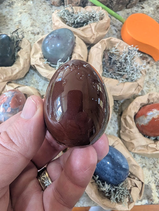 A 2-inch polished Sardonyx egg held in a hand, displaying a deep reddish-brown natural stone surface with a high-gloss finish.