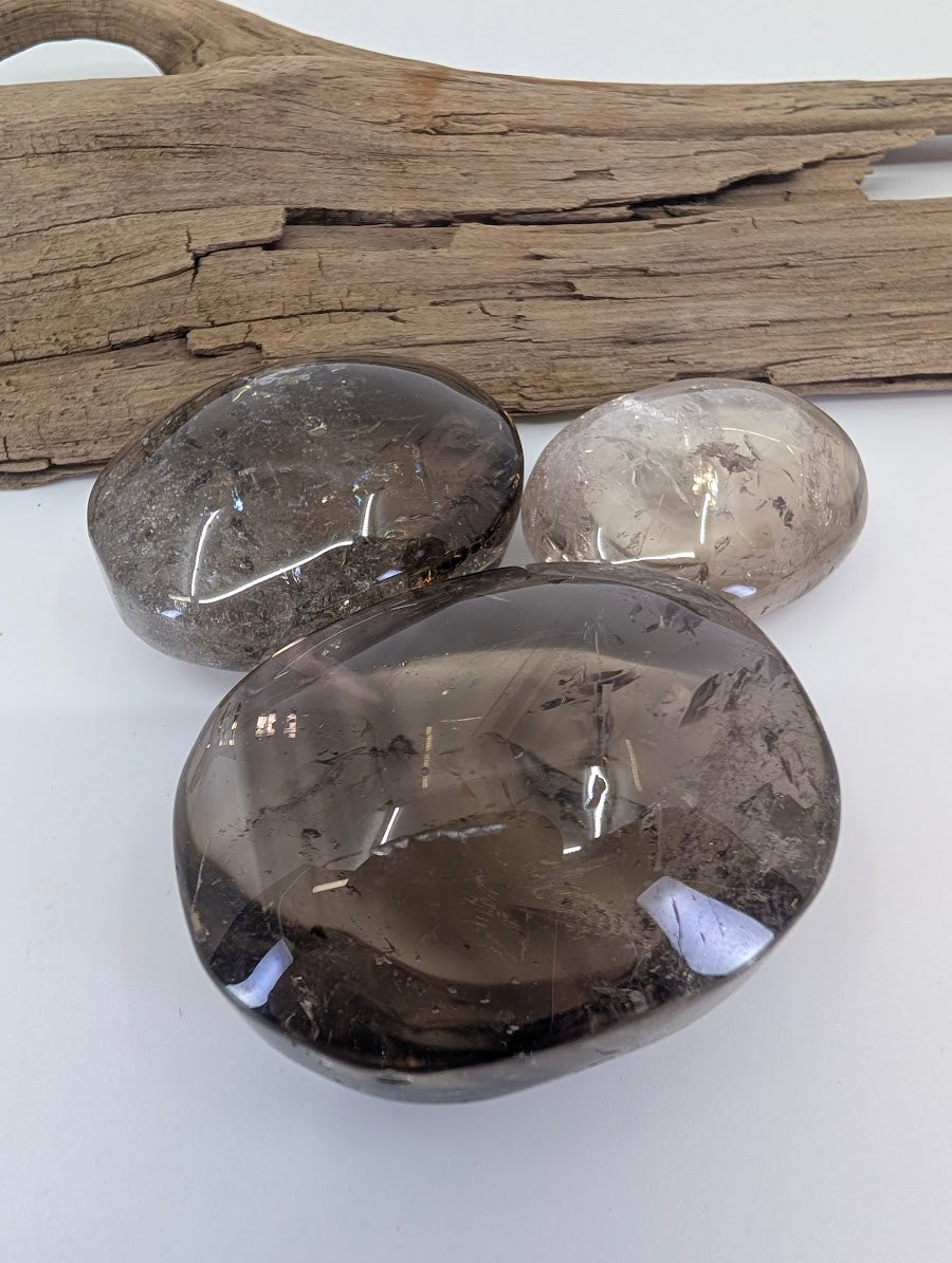 Trio of polished Lwena Smokey Citrine palm stones on a white surface with driftwood accent.