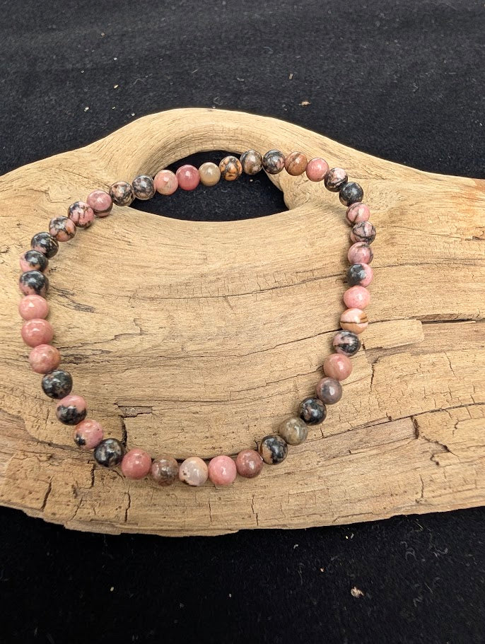 4mm polished Spider Web Rhodonite beaded stretch bracelet resting on a driftwood display, showing rose pink tones and black veining.