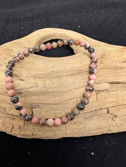 4mm polished Spider Web Rhodonite beaded stretch bracelet resting on a driftwood display, showing rose pink tones and black veining.