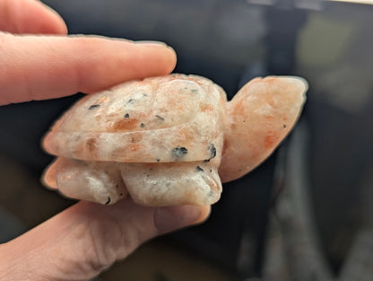 Side profile of a polished 60mm Sunstone turtle carving held between fingers, displaying peachy tones and natural dark mineral inclusions.
