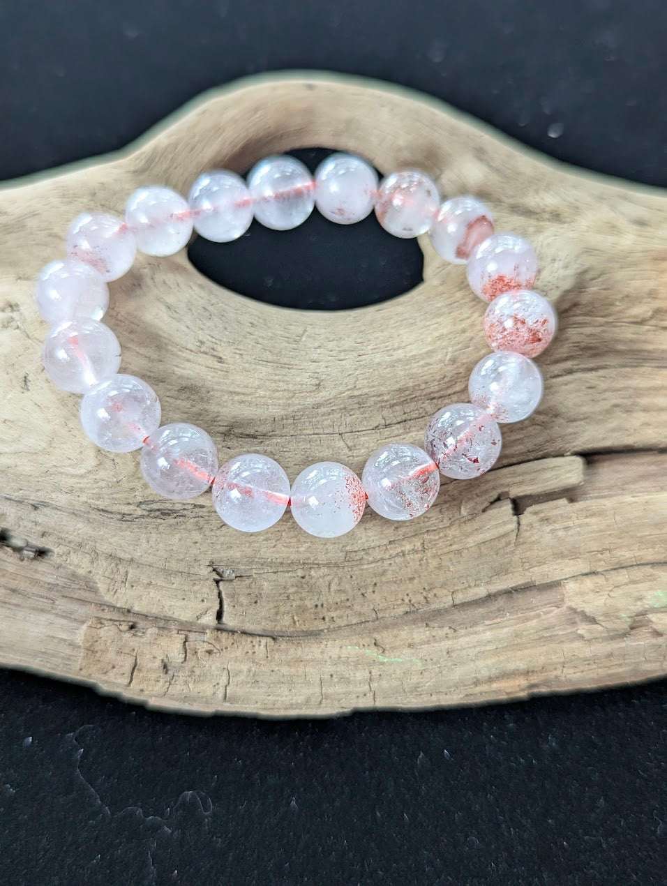 Large 10mm Super Seven bracelet displayed on natural driftwood, showing excellent bead clarity and deep red mineral deposits