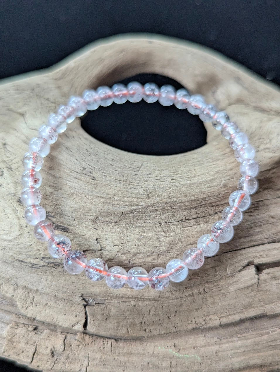 5mm AA Grade Super Seven gemstone bracelet from the Congo resting on natural driftwood, showcasing clear quartz with red lepidocrocite inclusions.