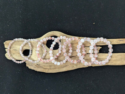 A full lineup of Super Seven bracelets from the Congo ranging from 5mm to 11mm sizes, arranged sequentially on natural driftwood.