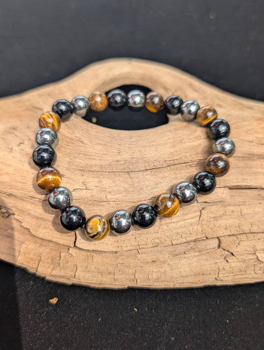 An 8mm Triple Protection beaded bracelet resting on natural driftwood, displaying golden Tiger Eye, metallic Hematite, and Black Obsidian.