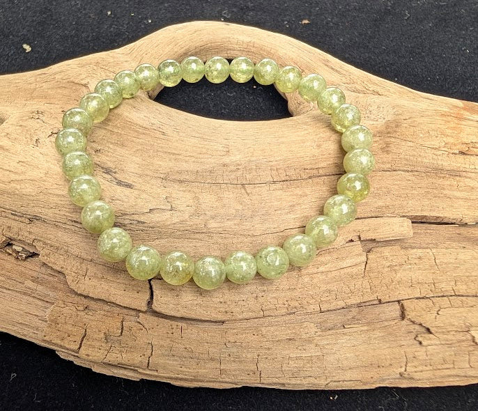 7mm polished Tsavorite beaded stretch bracelet resting on a driftwood display, showing translucent pale green tones and internal veiling.