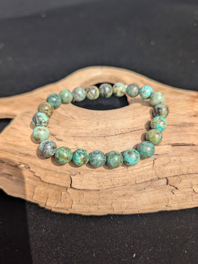 8mm natural turquoise beaded bracelet resting on a piece of driftwood, showing blue-green stones with dark matrix inclusions.