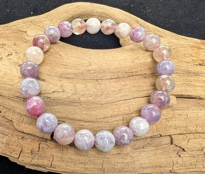 8mm polished Unicorn Stone pegmatite beaded stretch bracelet resting on a driftwood display, showing a mix of purple, pink, and translucent white minerals.