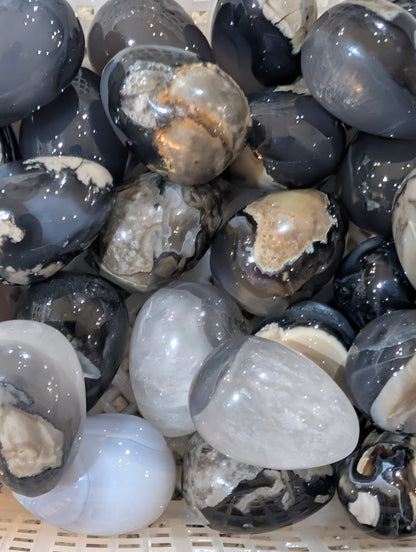 A collection of polished Volcano Agate eggs in a tray, showing variety in plume patterns and grey-scale tones.