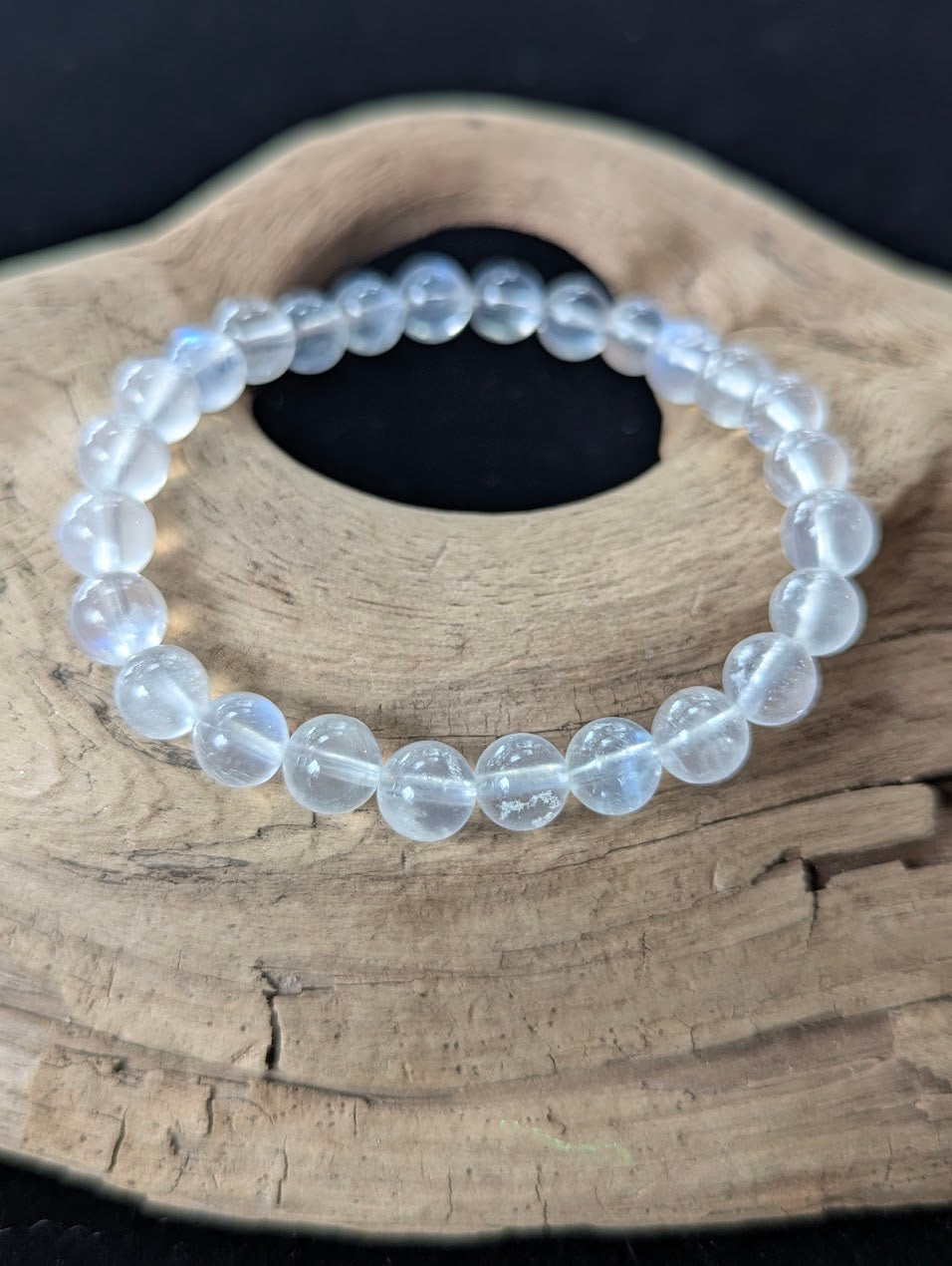 7mm AAA Grade White Moonstone gemstone bracelet resting on natural driftwood, showcasing high-clarity translucent beads with blue adularescence.