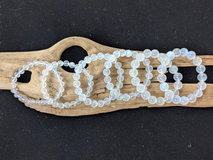 A full lineup of AAA Grade Sri Lankan White Moonstone bracelets ranging from 7mm to 12mm sizes, arranged sequentially on natural driftwood.