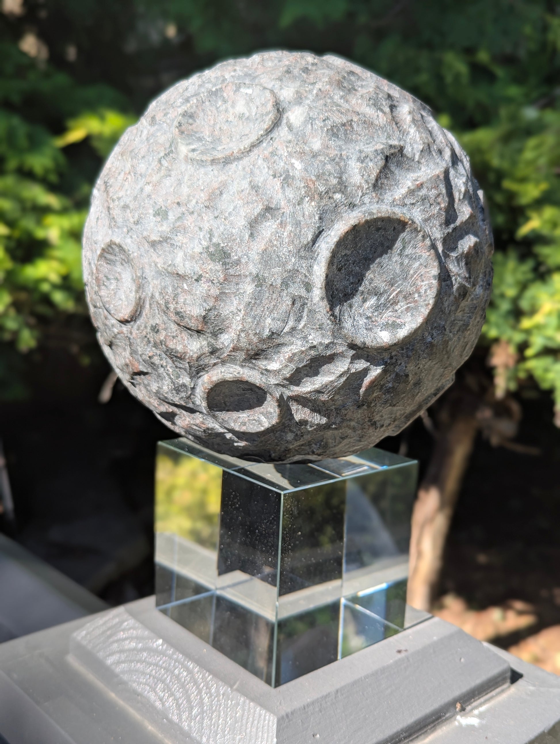 A 12 kg Yooperlite carved moon sphere in natural sunlight, showing its textured grey syenite matrix resting on a clear cubic stand.