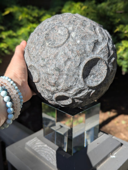 A hand holding a massive 210 mm (8.27 in) Yooperlite carved moon sphere, demonstrating its 12 kg scale and intricate crater carvings.